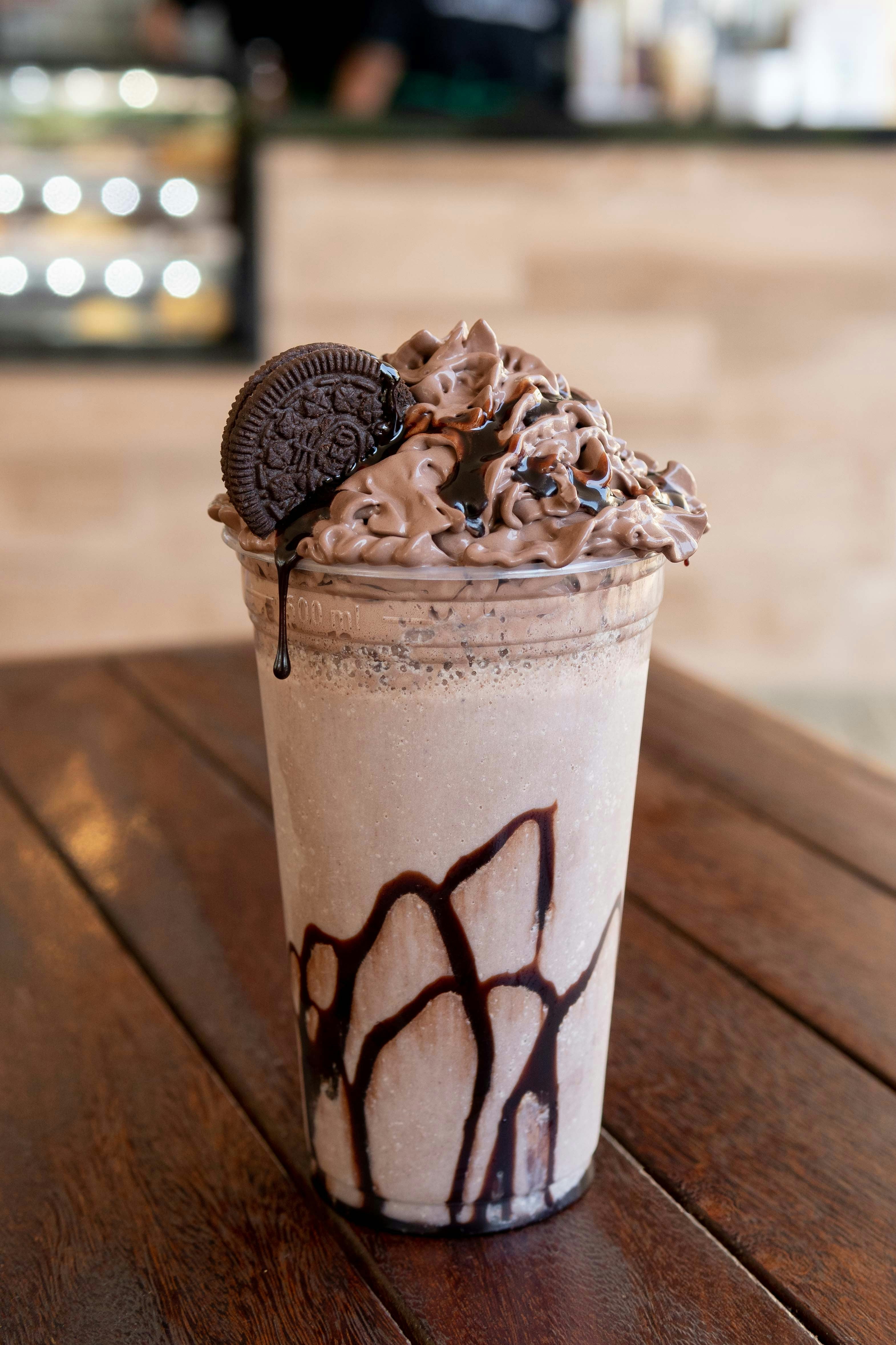 A tall glass of Frappuccino topped with creamy chocolate whip, drizzled chocolate syrup, and a cookie, sits on a wooden table. The background is blurred.