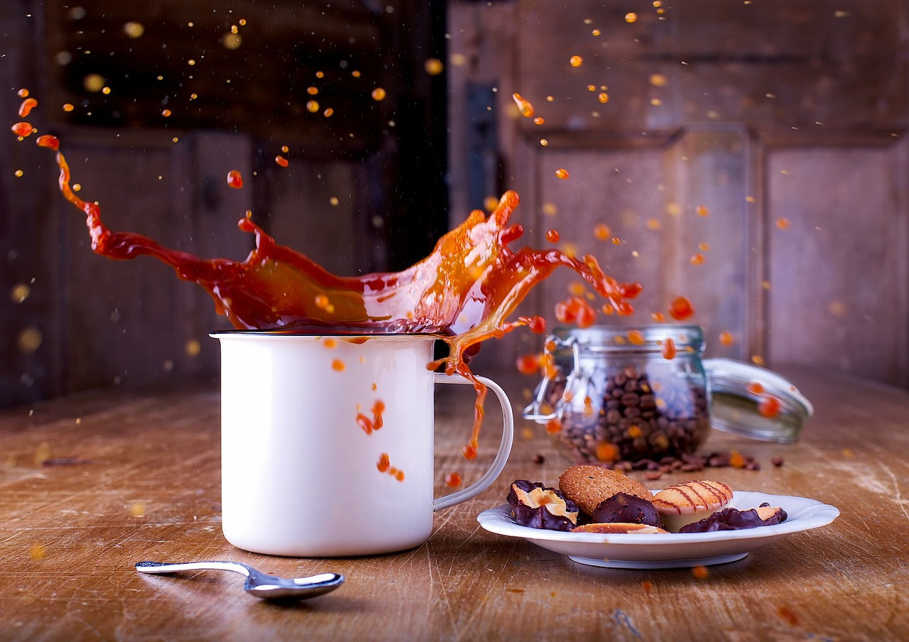 An overflowing white cup of coffee spills onto a wooden table, with a spoon nearby. A plate of cookies and a jar of coffee beans are in the background. The scene conveys a moment of lively chaos.