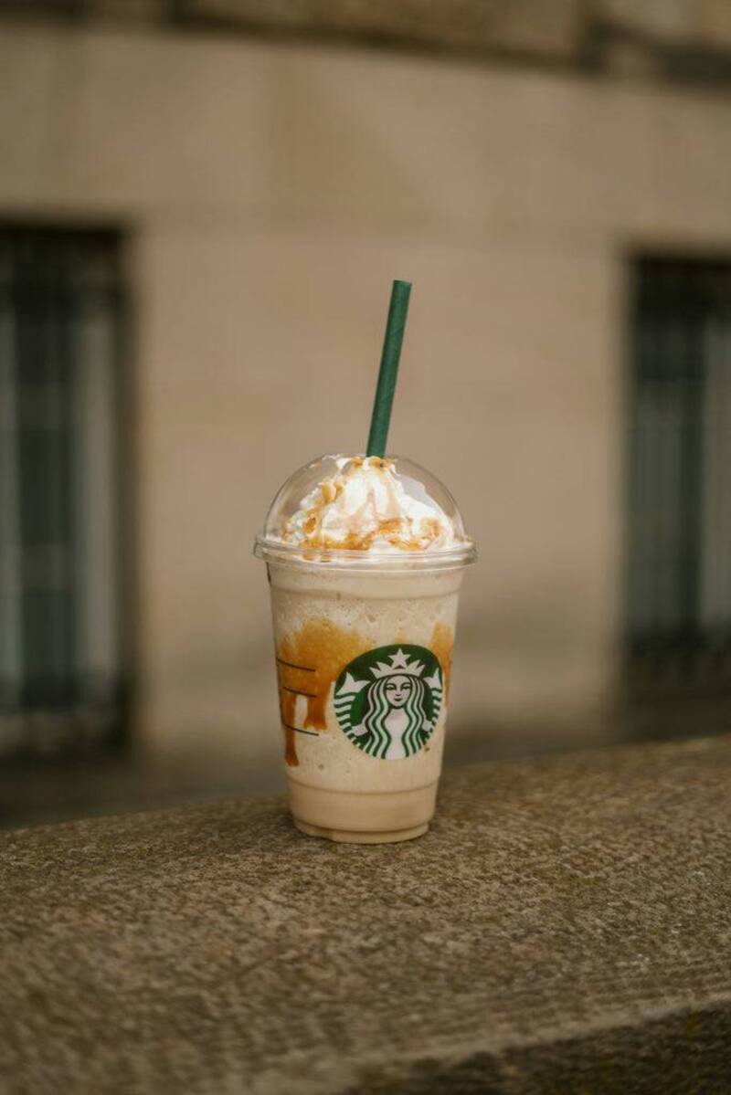 A Starbucks caramel frappuccino with whipped cream and drizzle, in a clear cup with a green straw, sits on a stone ledge.